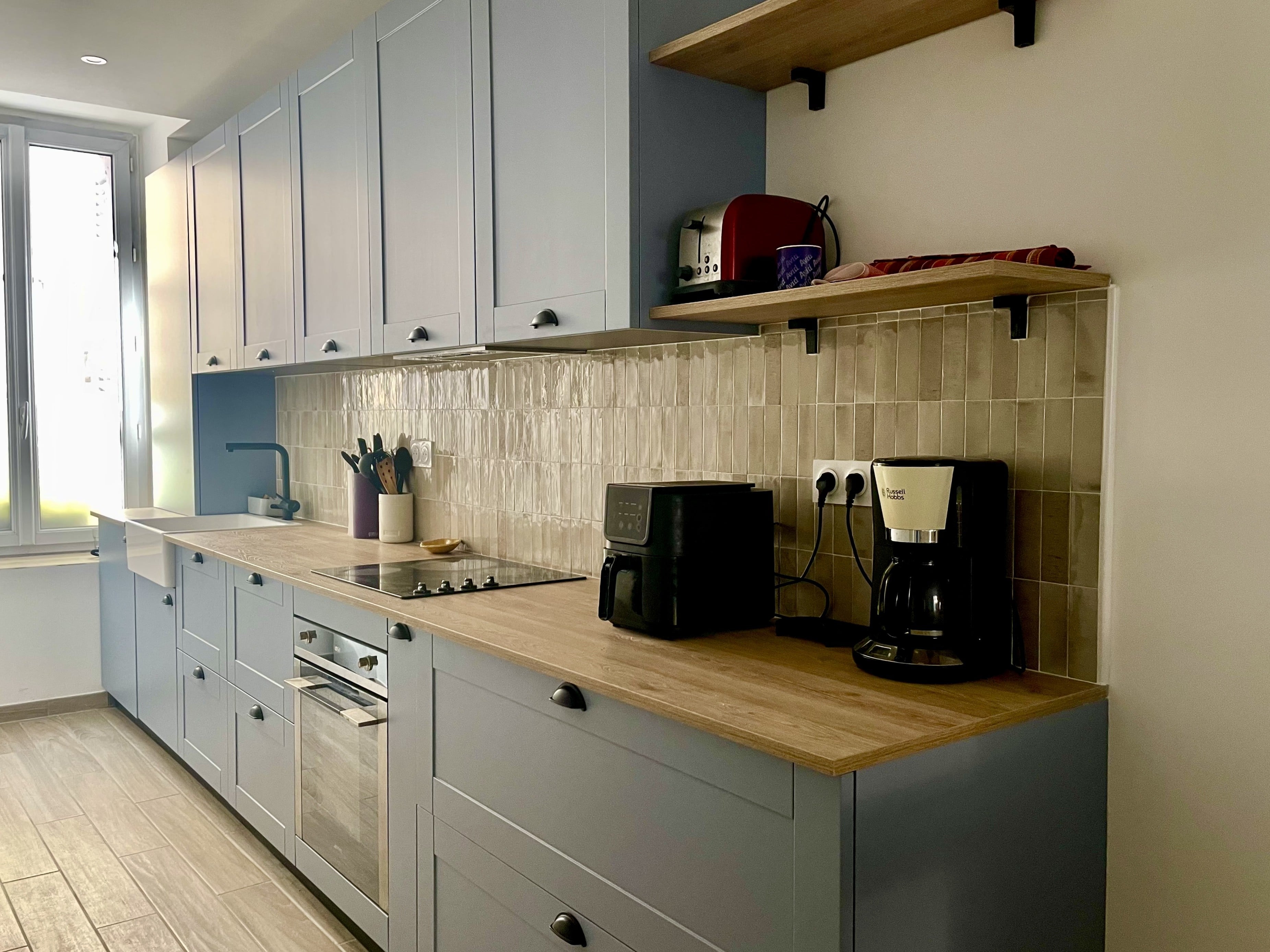 Cuisine linéaire aux façades à cadre bleu, plan de travail effet bois, crédence en zellige beige et étagères en bois