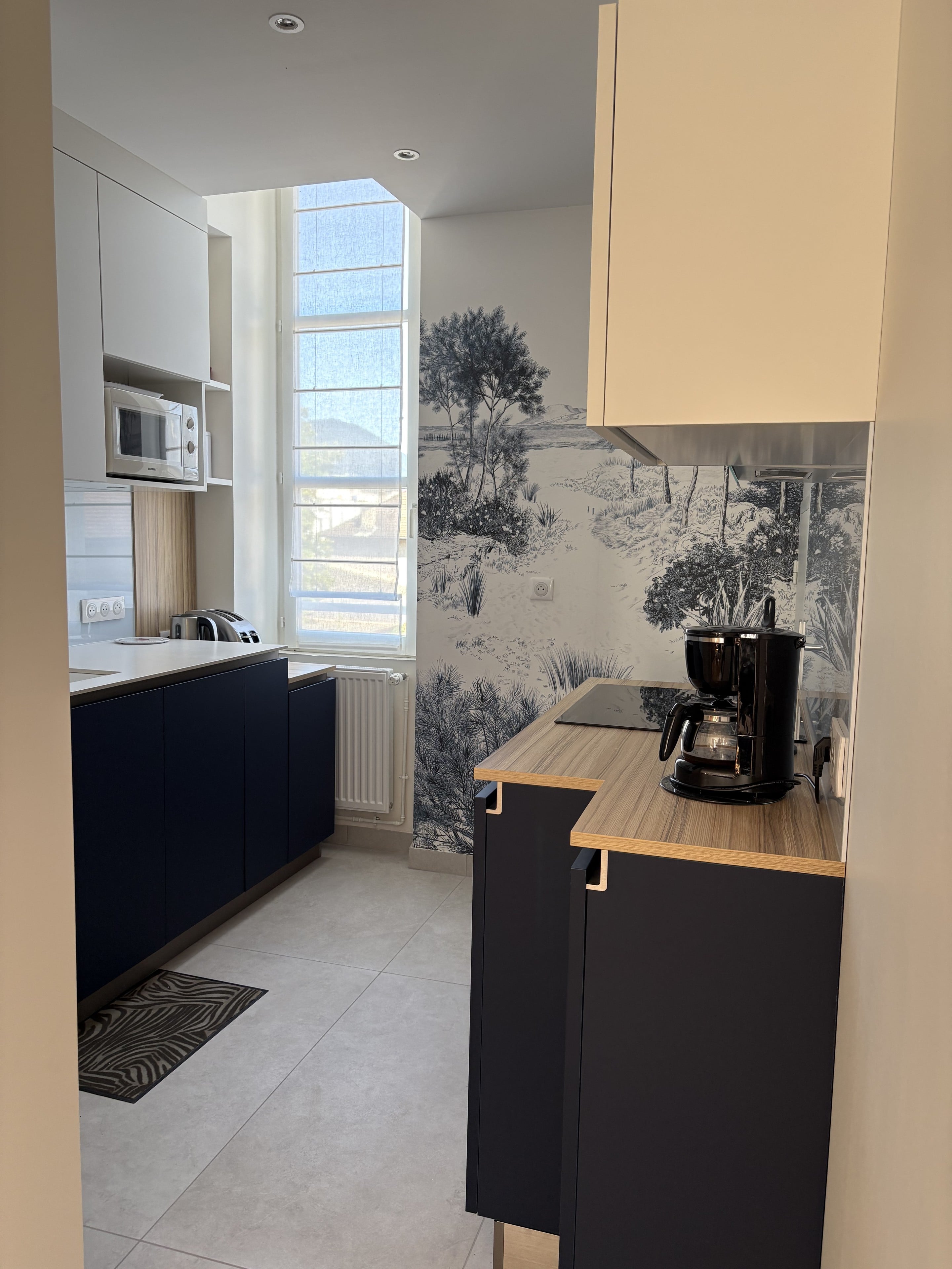 Cuisine en longueur aux meubles bas bleu nuit et hauts blancs, retour en plan effet bois et papier peint panoramique au fond