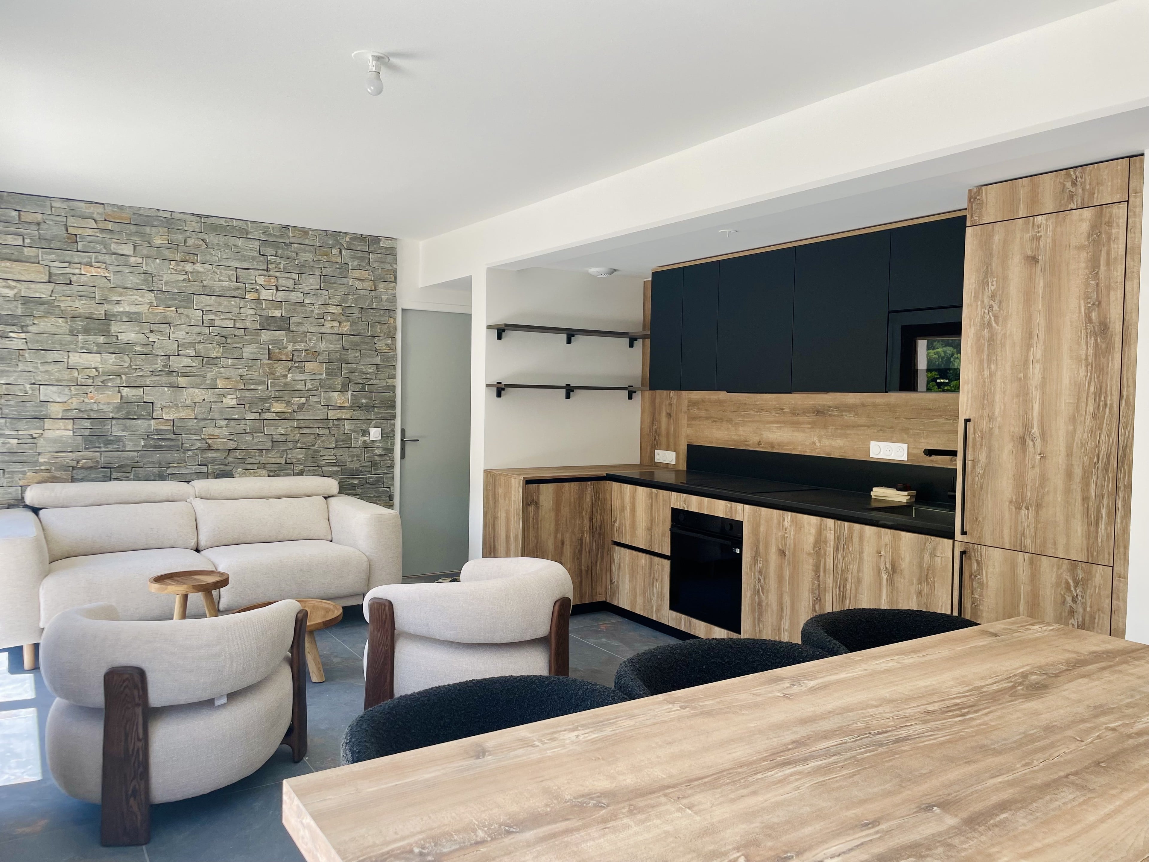 Pièce de vie ouverte : cuisine en chêne et noir mat au fond, mur en pierre, fauteuils bouclés et table en chêne au premier plan