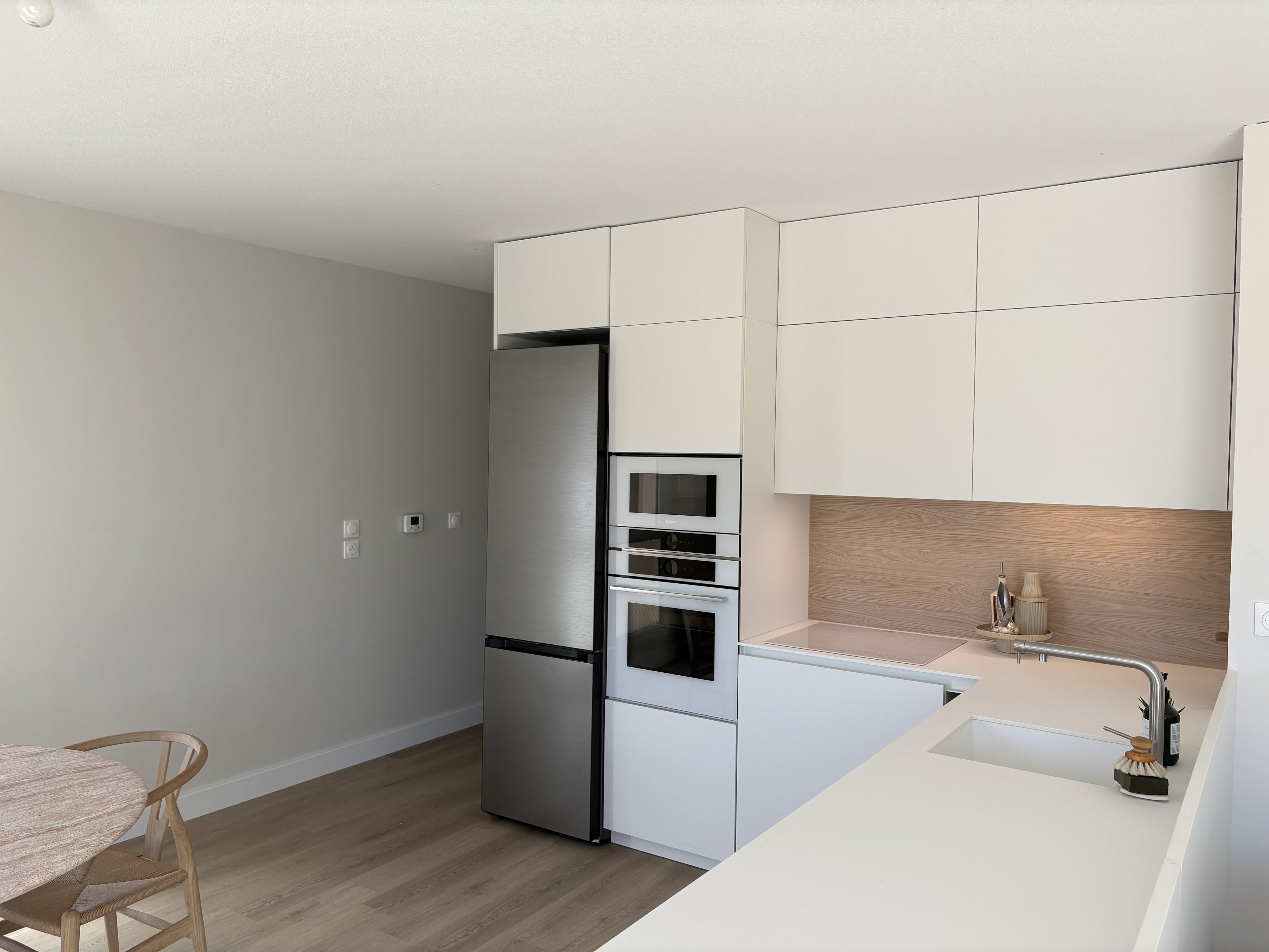 Cuisine blanche sans poignées, crédence effet chêne éclairée sous les meubles hauts, évier sous plan dans le retour