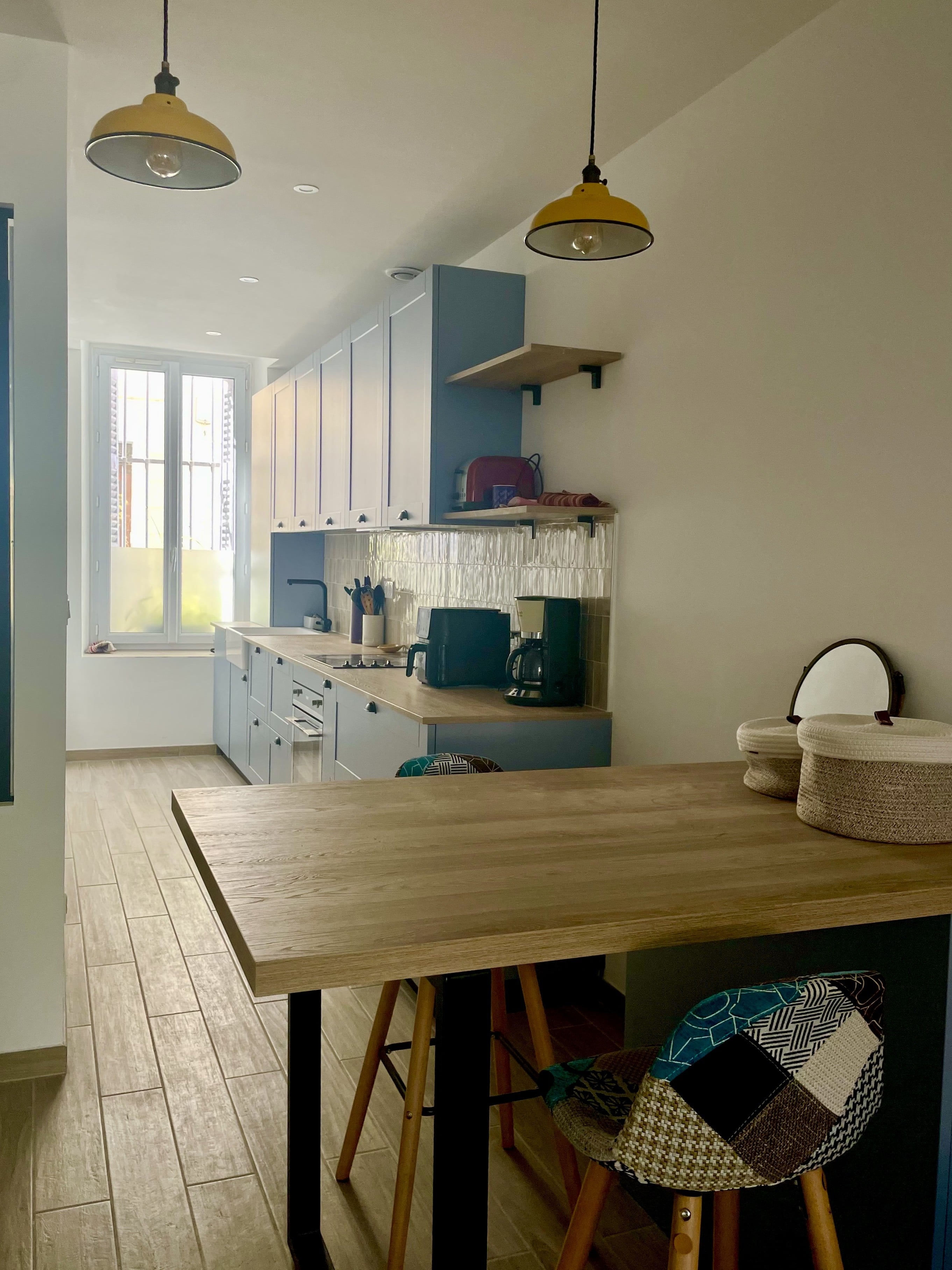 Cuisine en longueur aux façades bleues à cadre, table haute effet bois et suspensions jaunes, fenêtre au fond