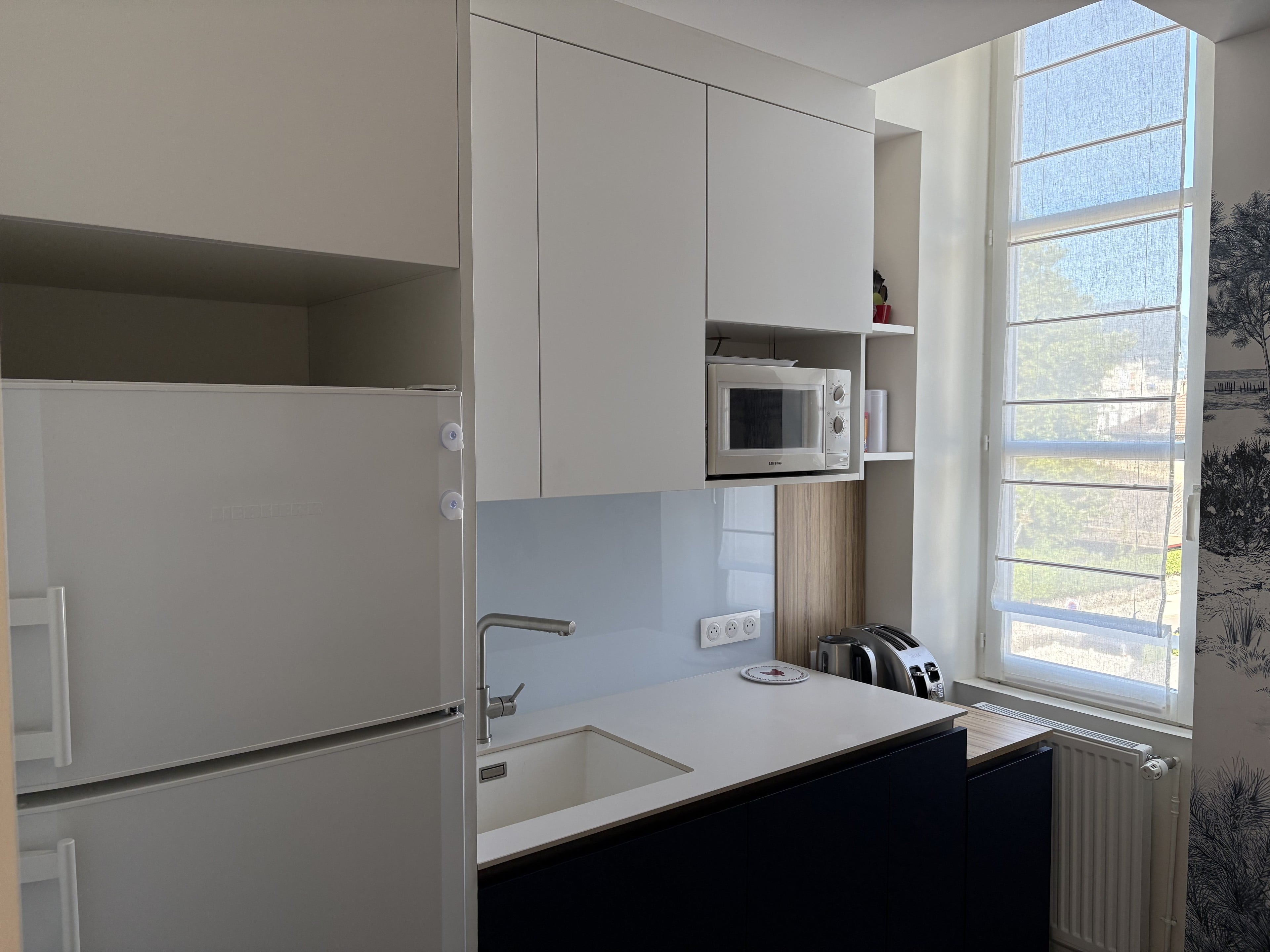 Meubles hauts blancs au-dessus de l’évier, haute fenêtre et papier peint panoramique, réfrigérateur blanc à gauche