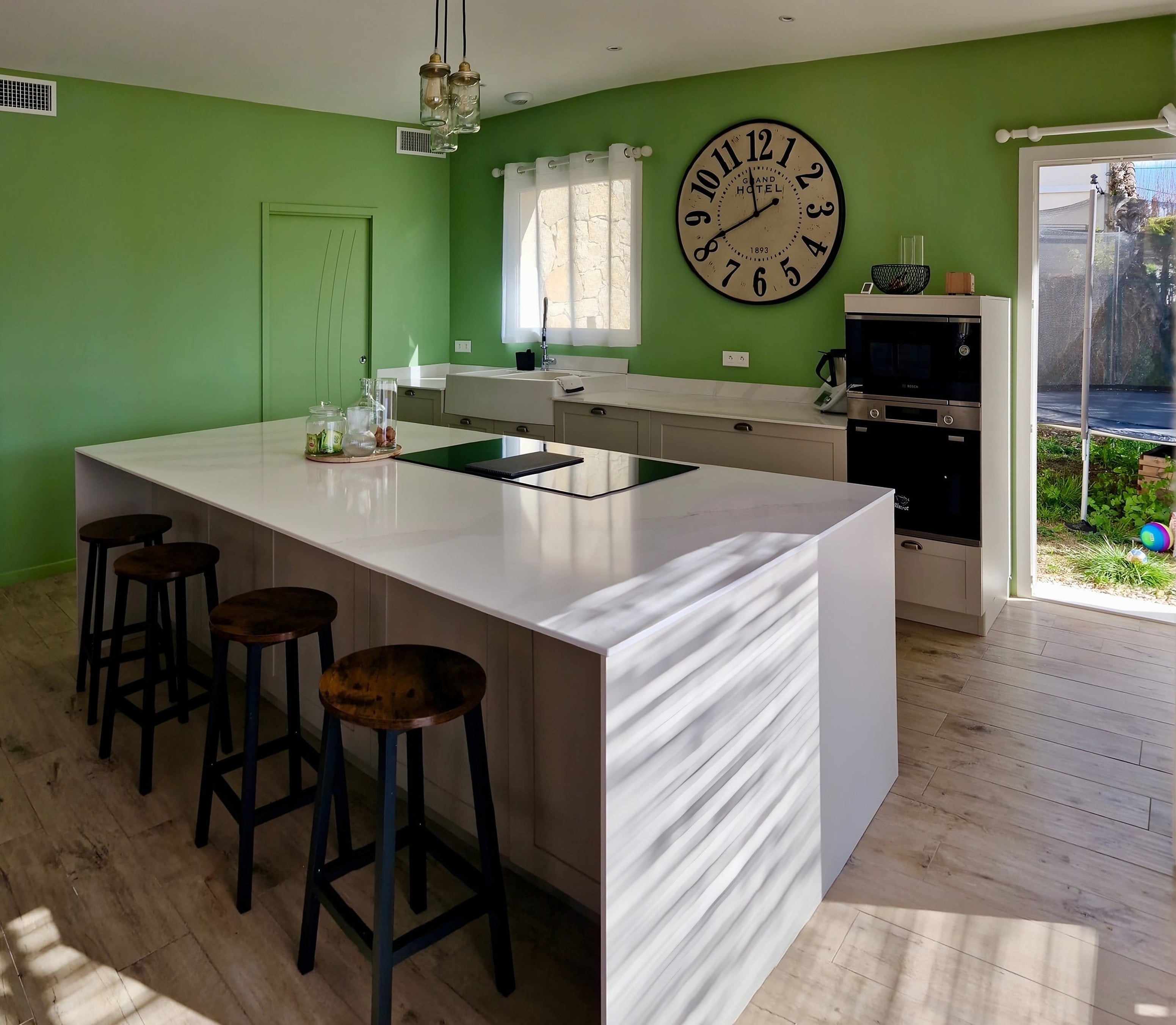 Grand îlot en quartz Calacatta Gold sans hotte grâce à la plaque aspirante, mur vert et tabourets en bois